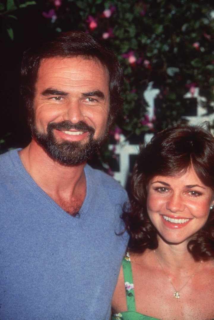 American actor Burt Reynolds smiles with his girlfriend, actor Sally Field, at an outdoor event.  He wears a blue V-neck sweater, and has a beard and mustache.  |  Source: Getty Images